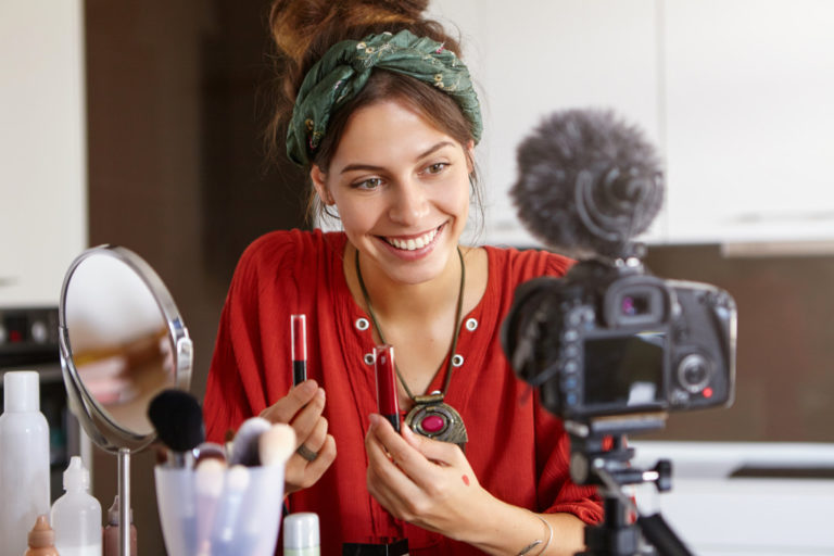 popular young woman video blogger advertising beauty products via her blog on social media, holding two lip gloss, comparing their quality, looking at camera fixed on tripod